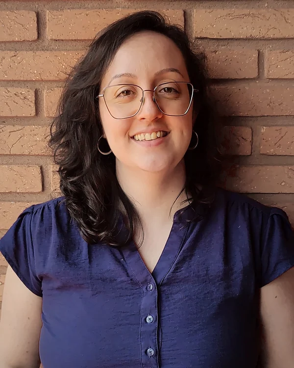 Retrato de Marina Bassi, arquiteta, de óculos e blusa azul, diante de uma parede de tijolos
